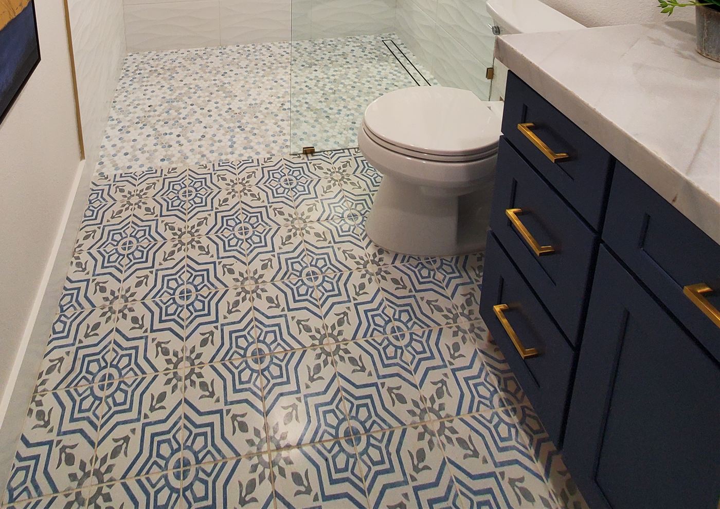 Bathroom floor in a blue-and-gray patterned tile with a small hexagon mosaic in the curbless shower, next to a navy vanity with gold pulls
